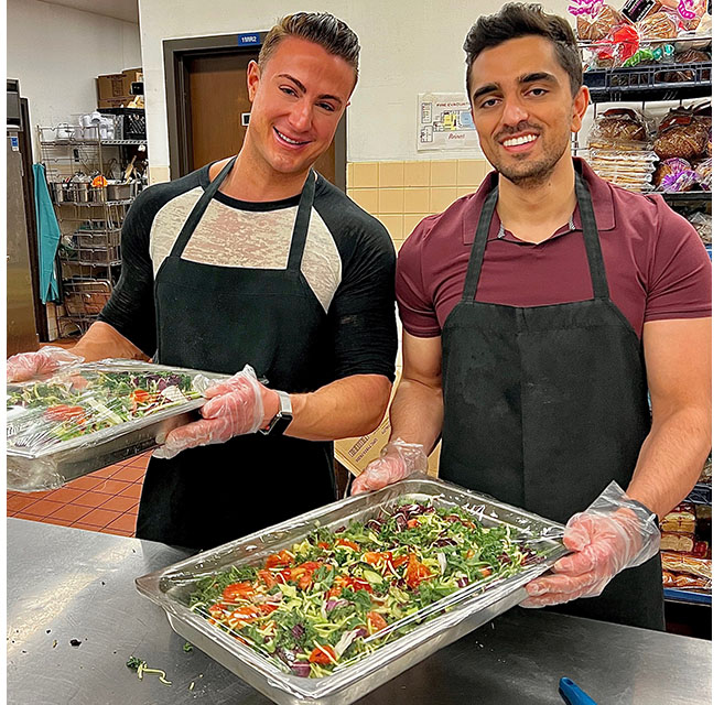 Two volunteers wearing aprons and gloves stand at a kitchen counter holding trays of salad.