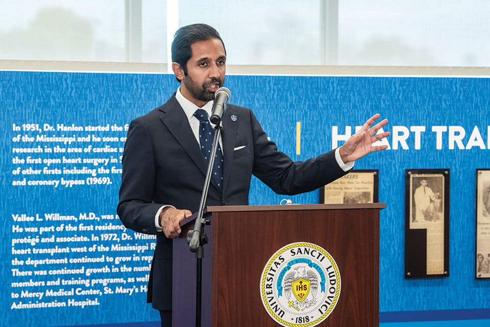 Sameer Siddiqui stands at a lectern in front of the surgery wall, speaking into a microphone.