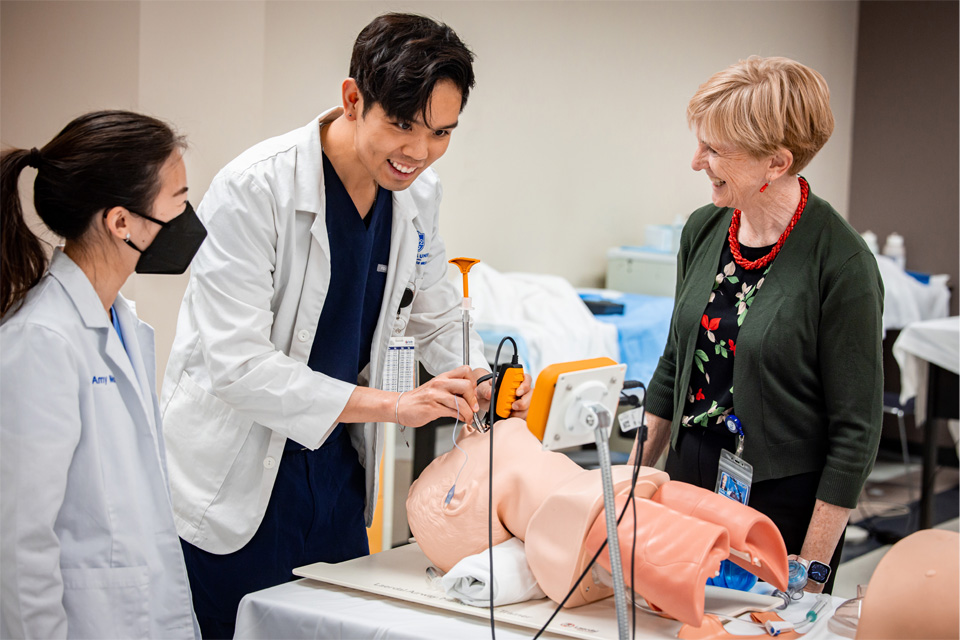 A medical student practices a procedure on a mannequin while a classmate and Chris Jacobs watch. All are smiling.