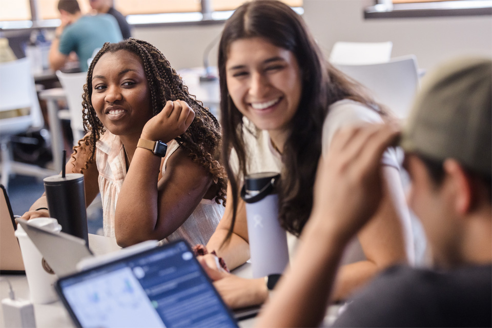 Two students laugh and smile while talking to a third who is working on a laptop.